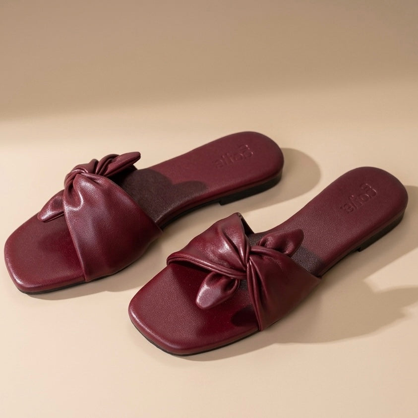 Pair of maroon sandals with bow details on a beige background